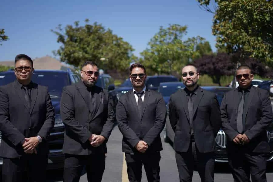 Five professional chauffeurs in black suits and sunglasses standing in front of luxury black SUVs for premium transportation services.
